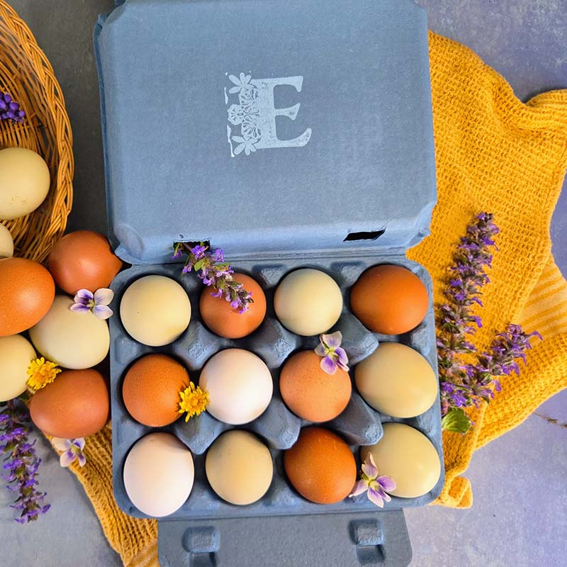 Carton of multicolored eggs with a decorative yellow cloth and flowers on a gray surface