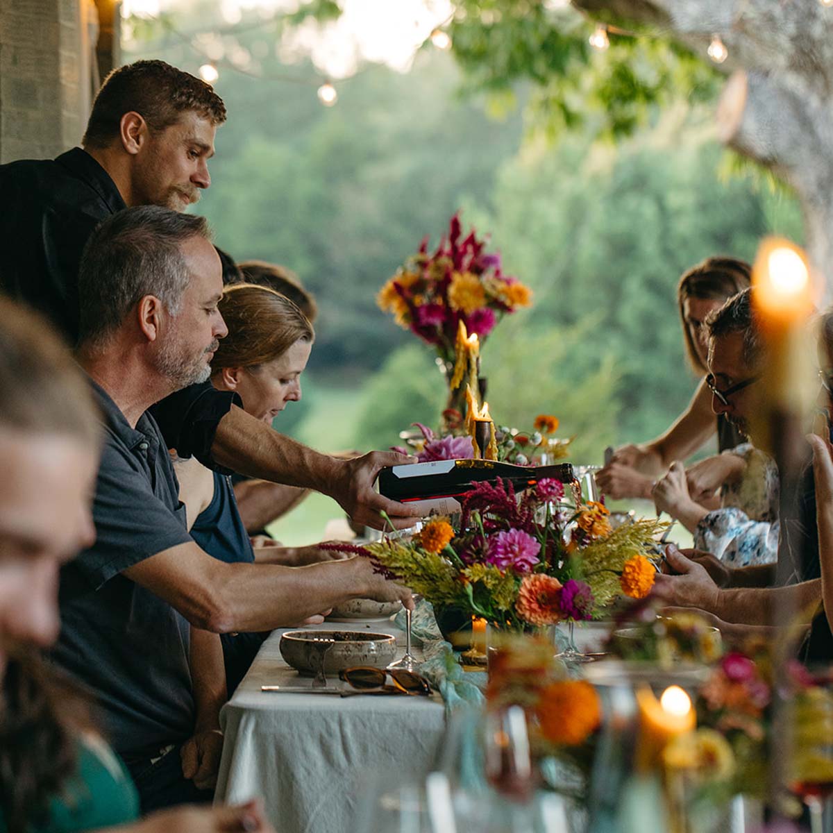 Group of people at a dinner table with floral arrangements and candles, enjoying a meal outdoors.