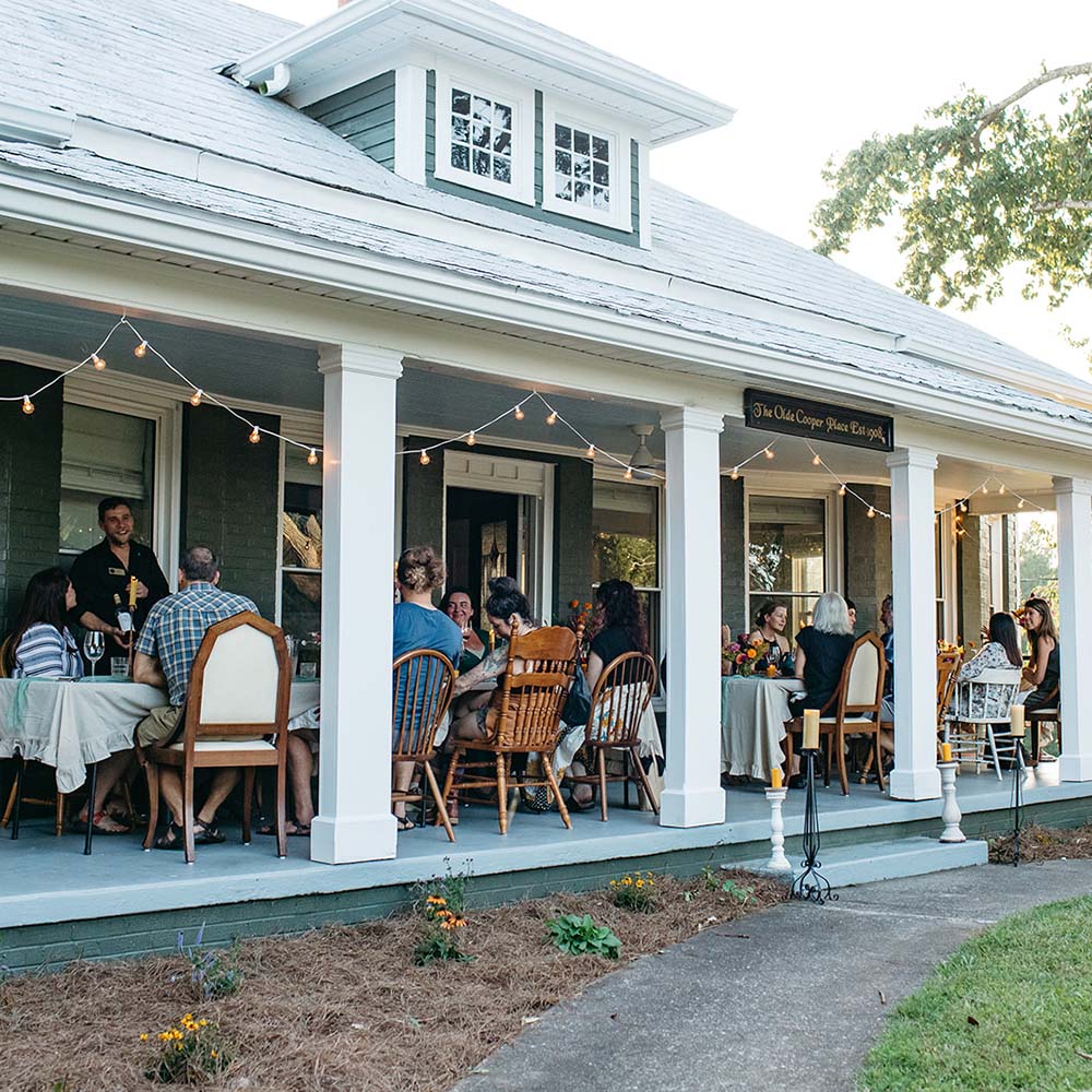 People dining on a porch with string lights and a white house in the background