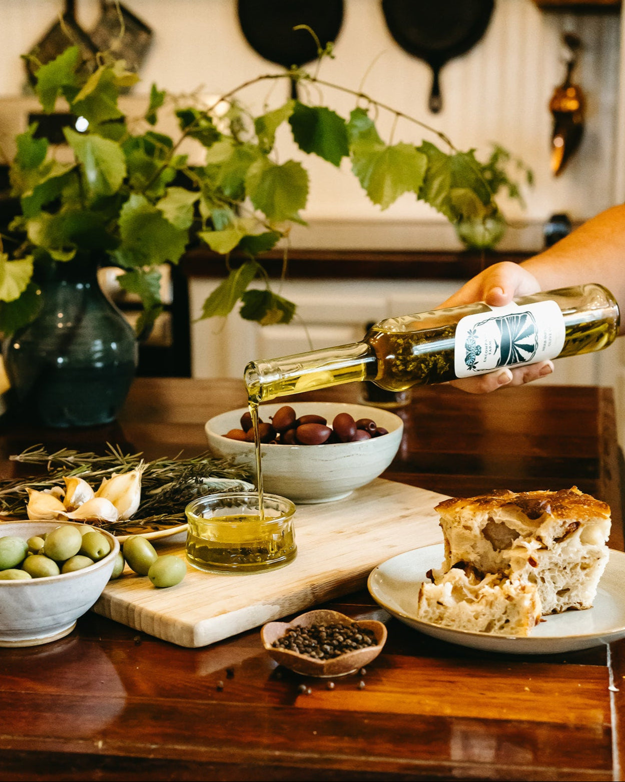 Pouring olive oil over a dish of food on a wooden table with greenery and kitchenware in the background.