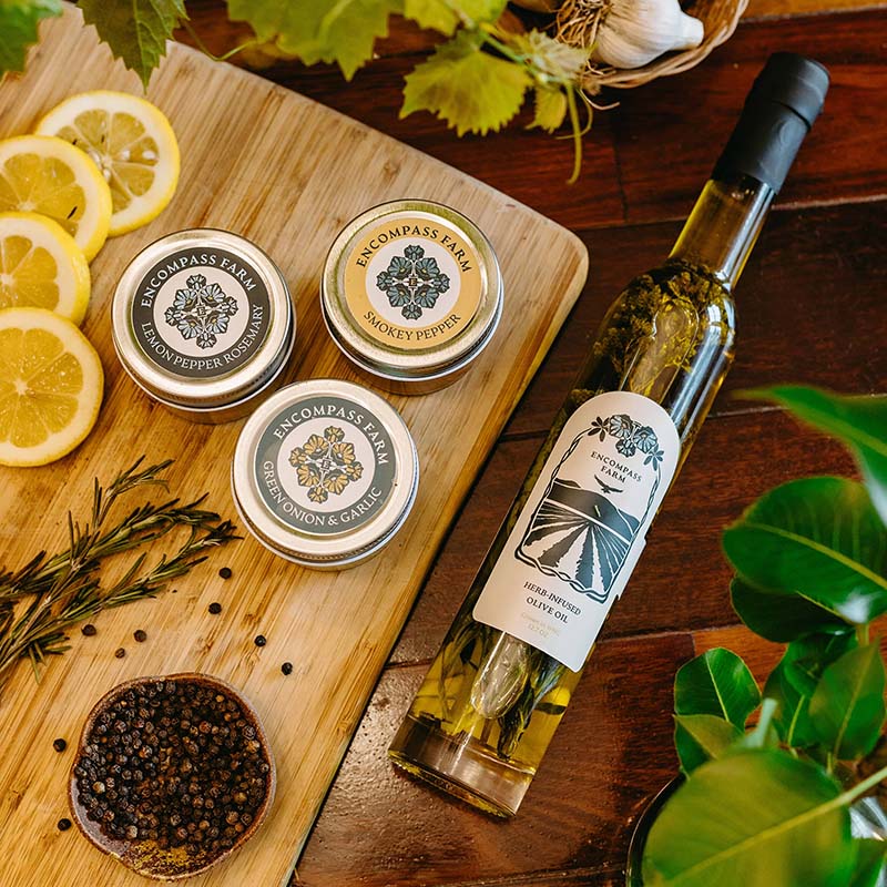 Bottle of Encompass Farm infused olive oil and jars of seasoning blends on a wooden board with lemon slices and herbs.