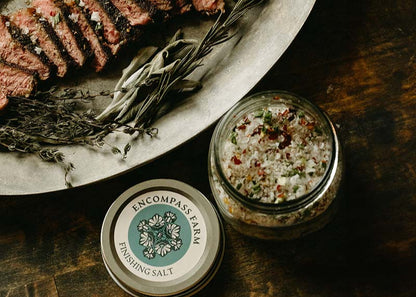Sliced steak on a platter with herbs and a jar of Encompass Farm finishing salt on a wooden surface