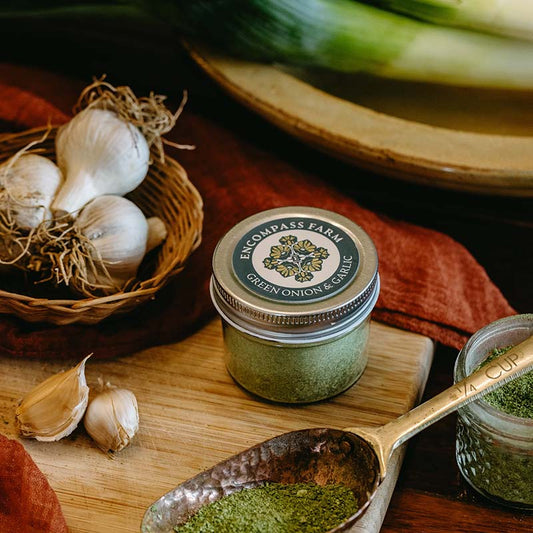 Jar of green onion and garlic seasoning from Encompass Farm on a wooden surface with garlic and green onions.