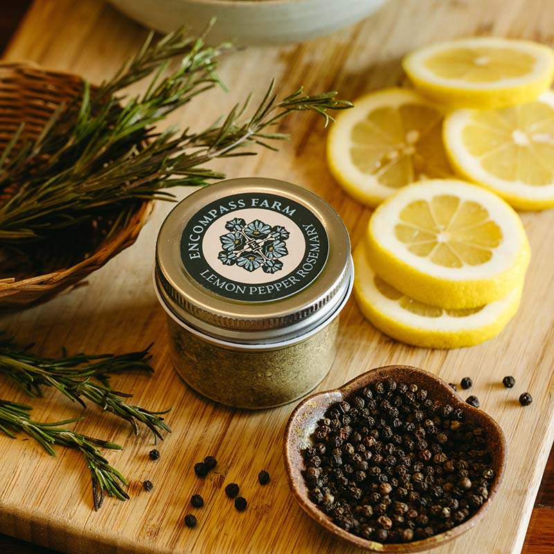 Jar of lemon pepper seasoning from Encompass Farm on a wooden cutting board with lemons, pepper and rosemary.