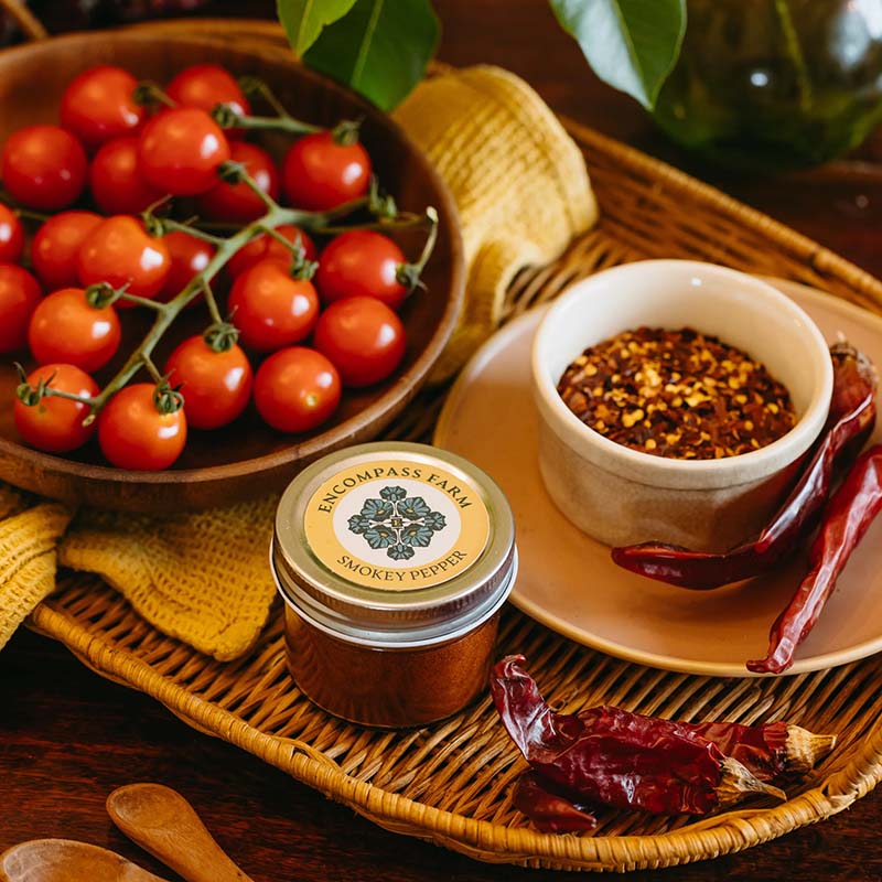 Jar of 'Encompass Farm Smoky Pepper seasoning' with cherry tomatoes and dried peppers on a woven tray.